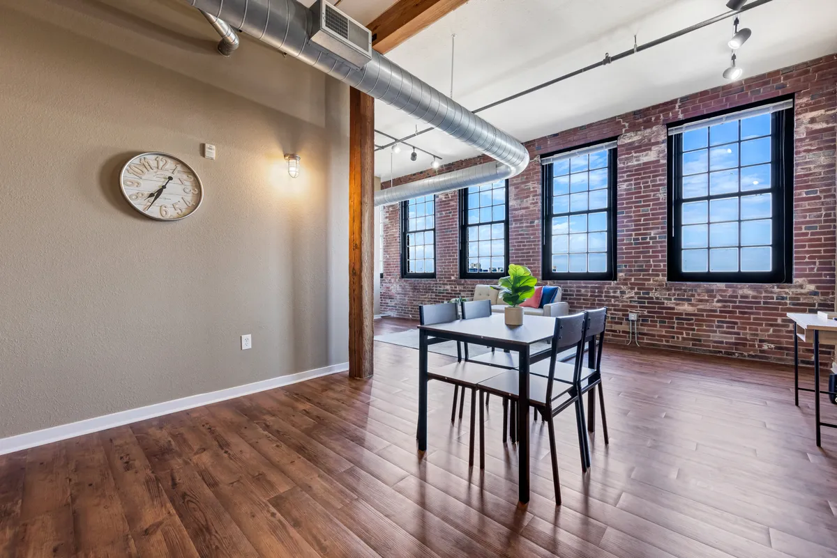 A spacious, light-filled dining area with a large table, exposed brick wall, and expansive windows. The space is versatile, accommodating not only dining and socializing but also quiet work or planning sessions, as indicated by the activities suggested. The mood is modern and inviting, perfect for both productivity and relaxation.