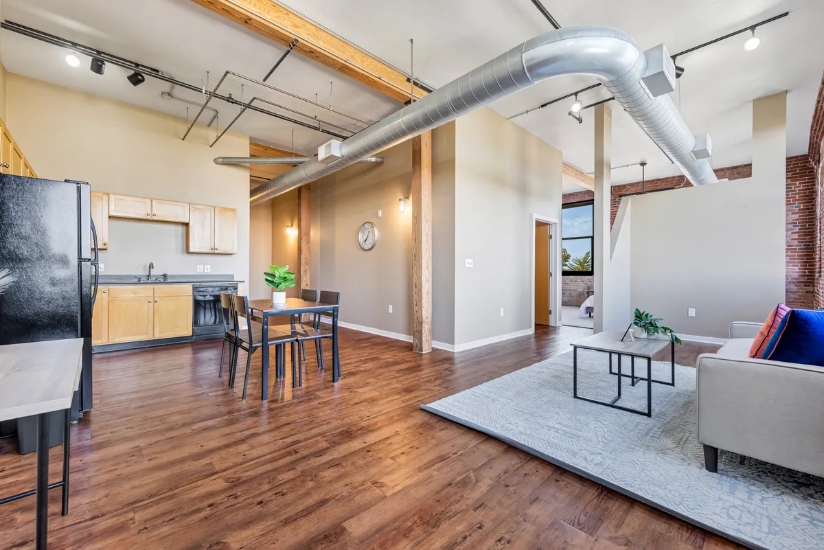 The image reveals an open-plan space with living room seating, a dining table, and a kitchen area. Exposed brick walls showcase the unit’s historic charm, while ample lighting and contemporary touches tie in the updated aesthetic. The seating and brickwork together highlight both comfort and character.