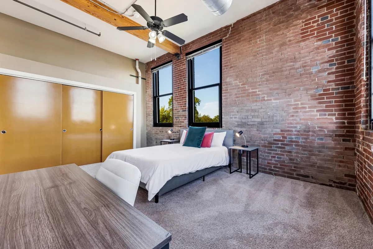 A cozy, modern bedroom featuring a plush bed against an exposed brick wall, large windows flooding the space with natural light, and a work desk for quiet moments. The overall mood is serene and comfortable.