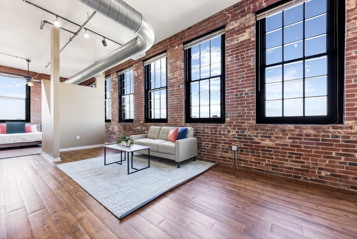This image features a spacious living area with a comfortable sofa, a coffee table, and a rug, all bathed in sunlight streaming through large windows. The modern furnishings are arranged to invite relaxation and conversation, while the scale of the windows emphasizes the room’s airy, open feel.