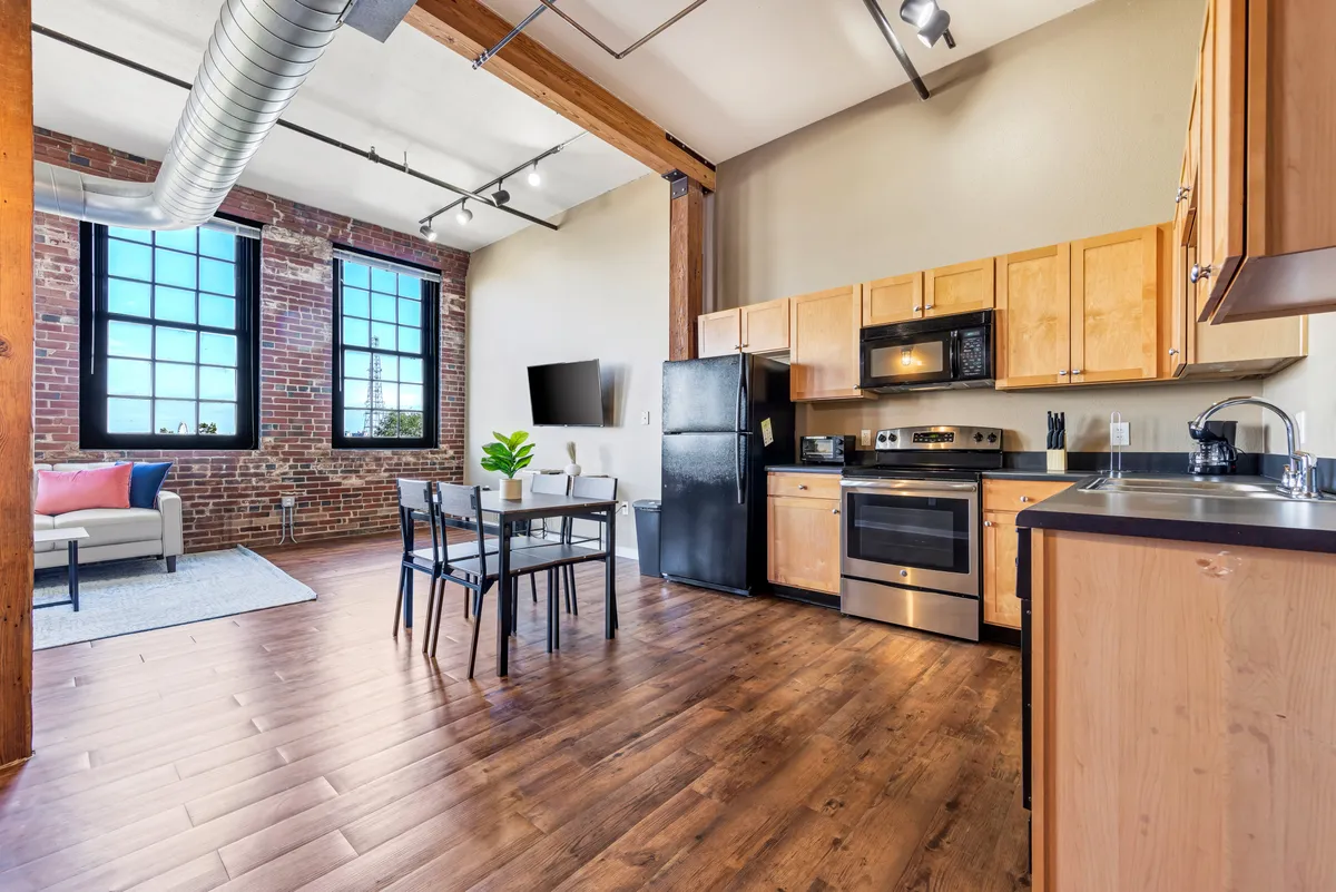An open-plan scene with large windows and exposed brick walls, showcasing a dining table adjacent to the kitchen area. The kitchen appliances are visible, and the setting suggests a space for both cooking and gathering, filled with natural light and historic architectural features.