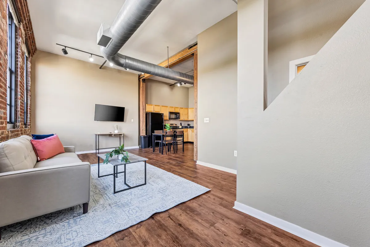 This image showcases a spacious living area featuring a comfortable sofa, coffee table, television, and an open kitchen area in the background. The room is inviting and well-lit, with a layout that suggests both relaxation and hosting potential. The high ceilings and large windows are visible, highlighting the modern comforts blended with historic charm.