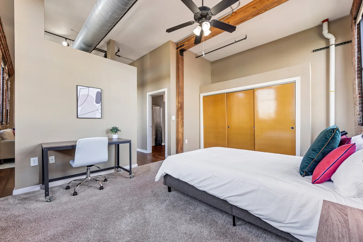 This image features a well-appointed bedroom with a comfortable bed, a work desk, a spacious closet, and eye-catching artwork on the walls. The scene is bathed in natural light, creating a cozy and stylish atmosphere.