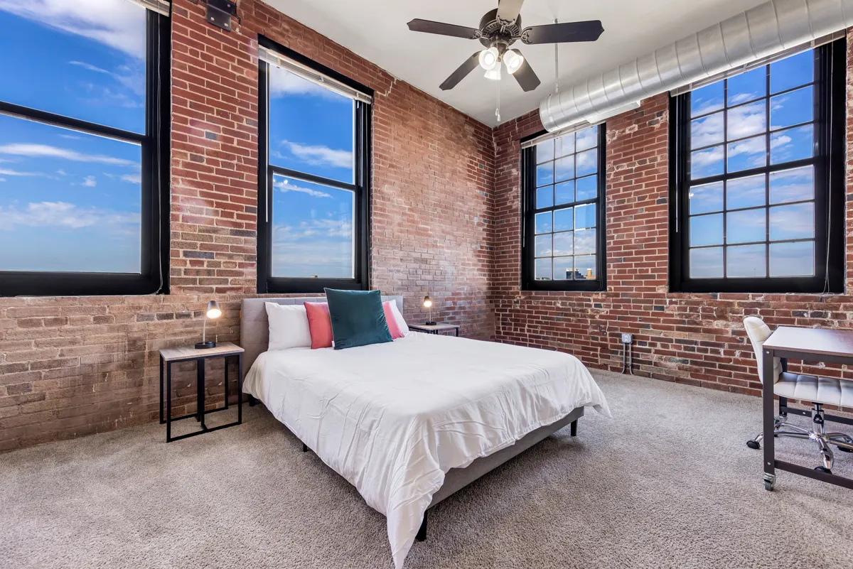 The image features a stylish bedroom with white bedding on a comfortable bed, set against exposed brick walls and large windows that flood the space with natural light. A small desk offers a spot for creative work or reading, and a ceiling fan adds comfort. The room blends historic charm and modern comfort, creating a cozy and inviting atmosphere.