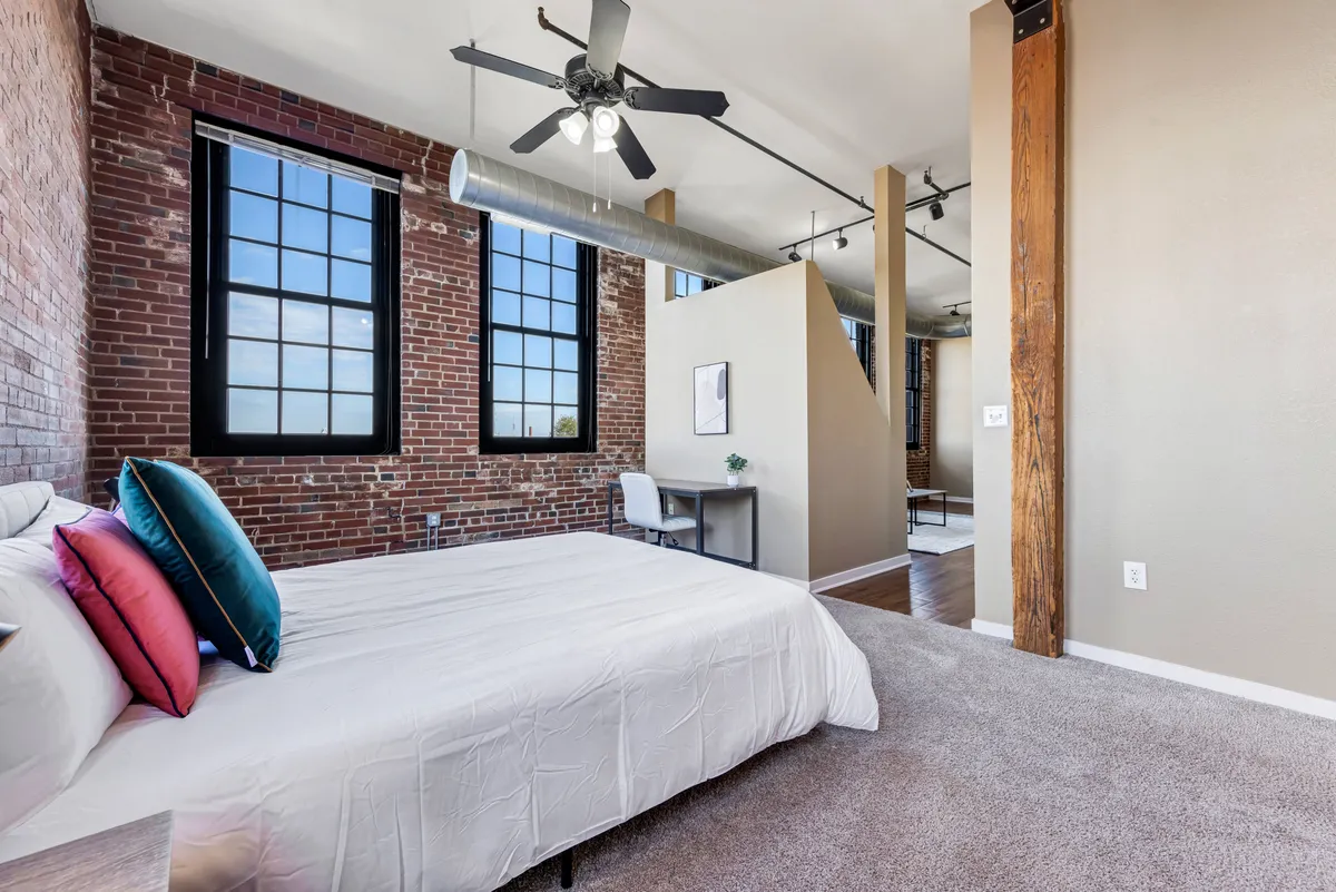 This image features a stylish interior with exposed brick walls, large windows streaming in natural light, modern bedding, and prominent wooden pillars. A person is depicted working at a dedicated desk, making use of the quiet, thoughtfully designed nook. The scene exudes a contemporary and cozy ambiance, perfectly tailored for productive work sessions.